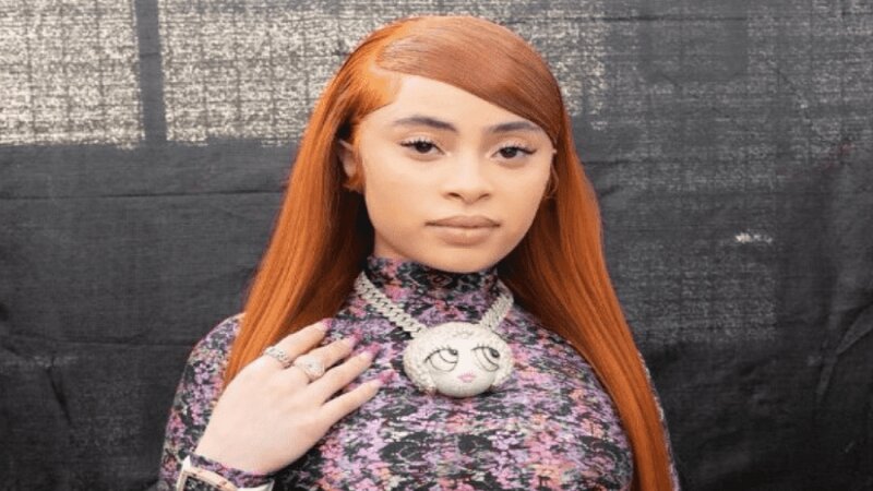 Portrait of Ice Spice with long orange hair, wearing a patterned high-neck top and a unique pendant necklace, standing against a dark backdrop