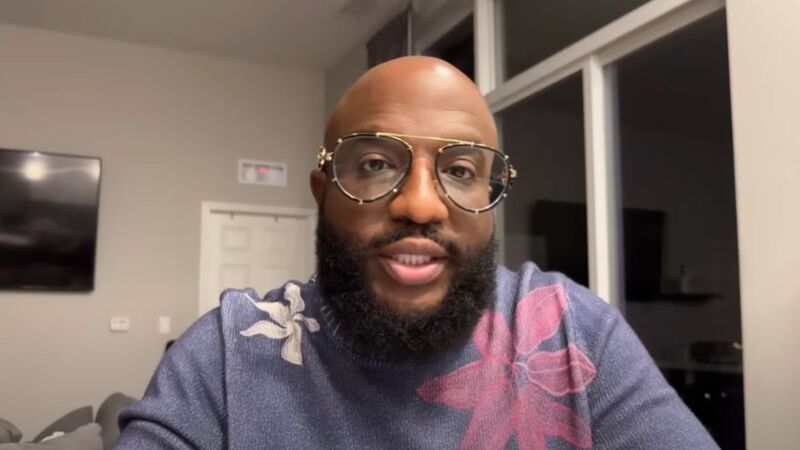 A man with a beard and glasses wearing a sweater with floral patterns, sitting indoors with a background showing a door and windows.