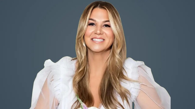 A smiling woman with long blonde hair, wearing a white ruffled blouse, against a plain blue background.